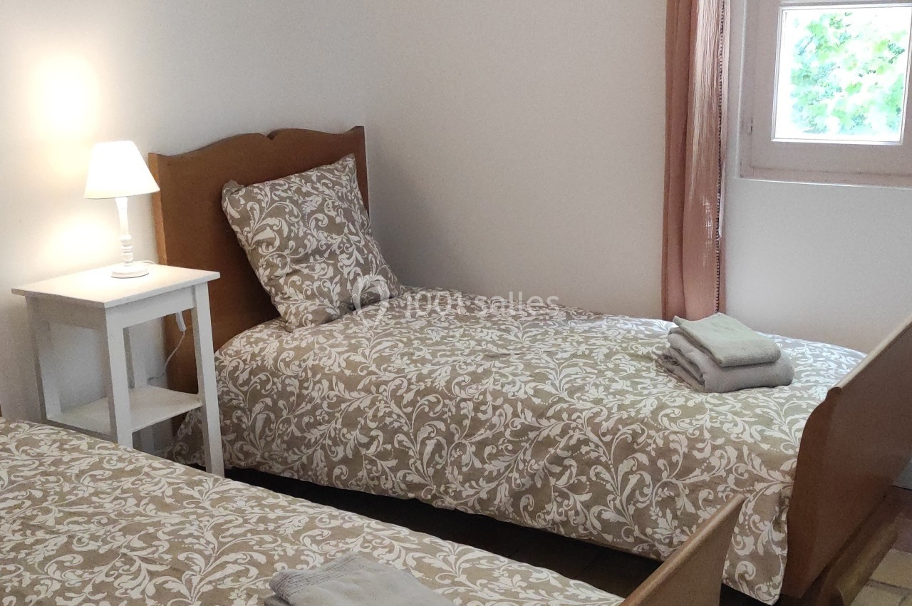 Deux lits simples avec linge de lit à motifs, une table de chevet avec lampe et une fenêtre laissant entrer la lumière.