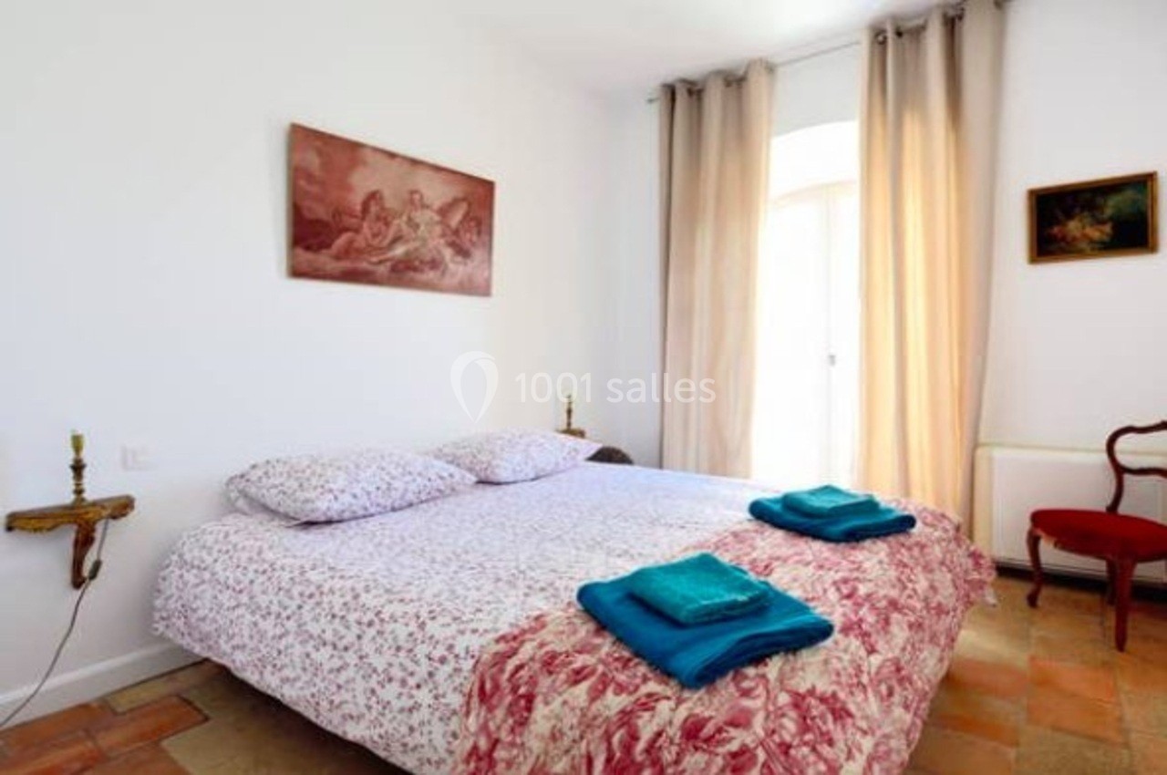 Lit double avec draps fleuris, deux serviettes pliées, murs blancs, rideaux beiges et tableaux décoratifs.