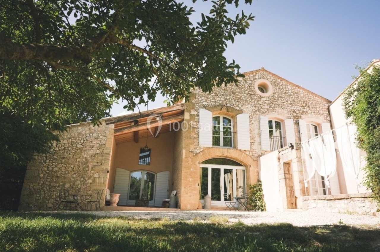 Façade d'une maison en pierre avec volets blancs, grande ouverture vitrée et terrasse ombragée par un arbre.