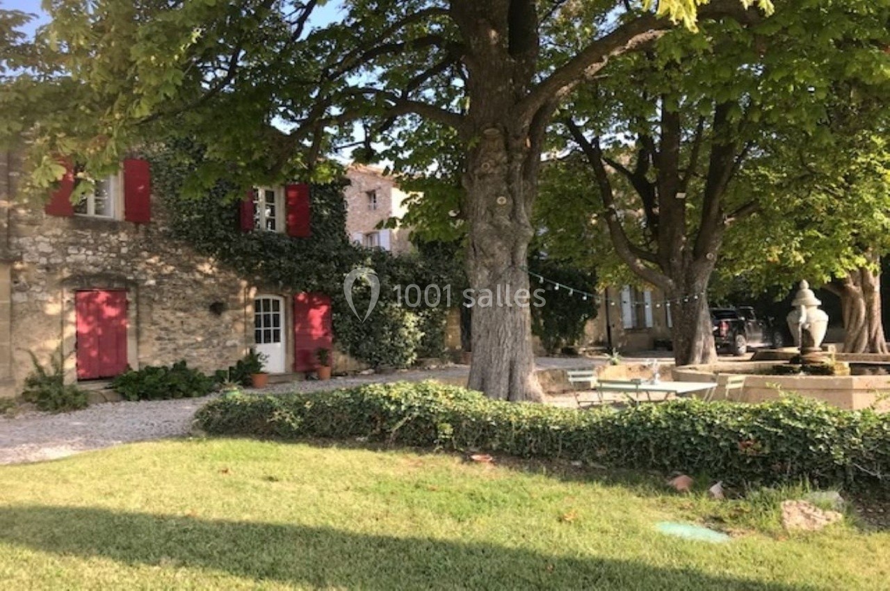 Maison en pierre avec volets rouges entourée d'arbres et de verdure, avec une cour aménagée et une fontaine.