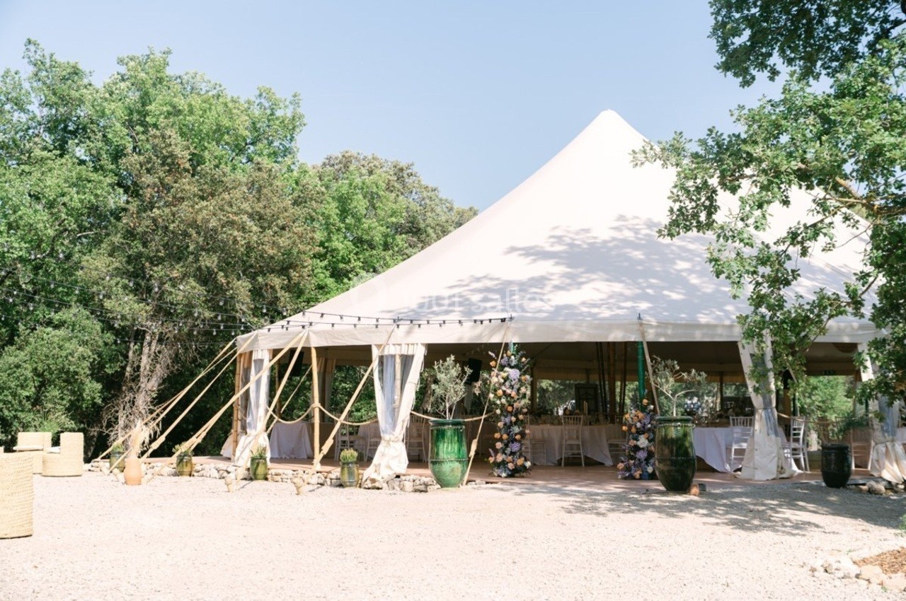 Grande tente blanche installée en extérieur, entourée d'arbres, avec des décorations et des tables visibles à l'intérieur.