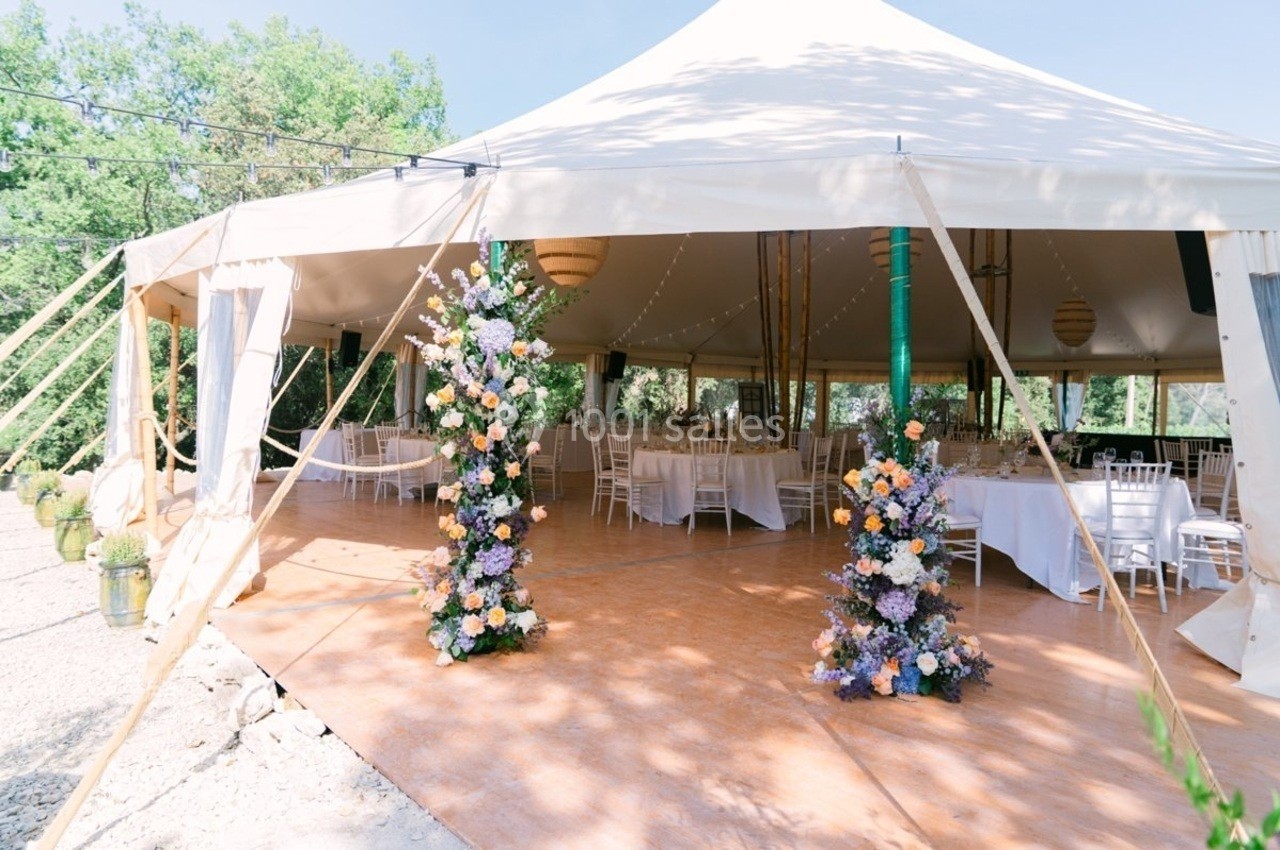 Tente blanche décorée de fleurs, abritant des tables et chaises pour un événement en extérieur par temps ensoleillé.