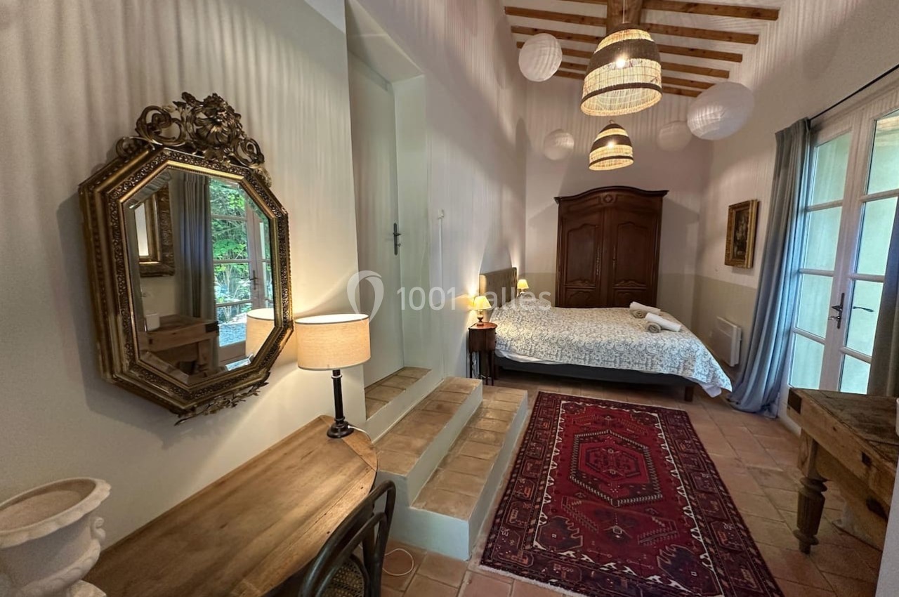 Chambre lumineuse avec lit double, bureau en bois, miroir orné, tapis rouge et plafond avec poutres apparentes.