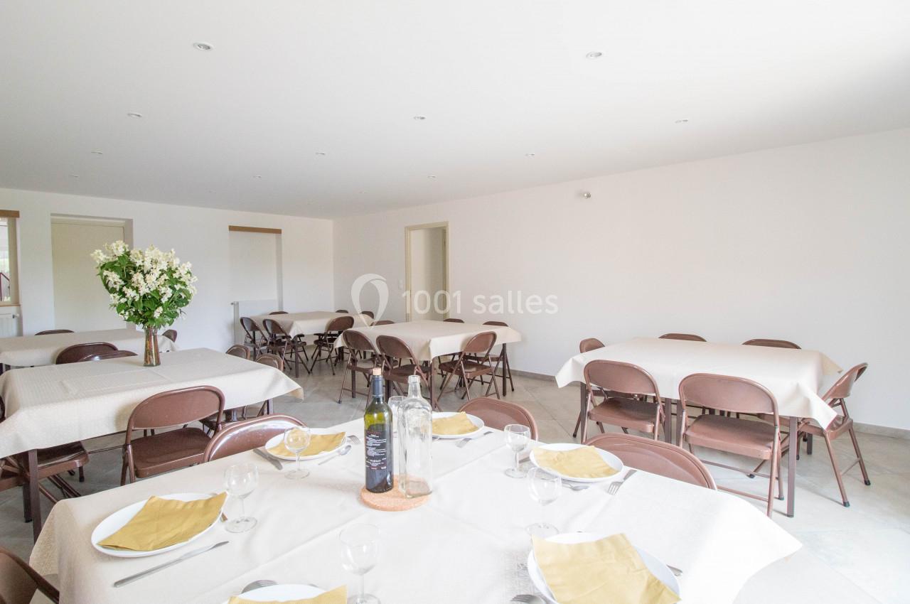 Salle lumineuse avec tables dressées, nappes blanches, chaises pliantes marron et un bouquet de fleurs en décoration.