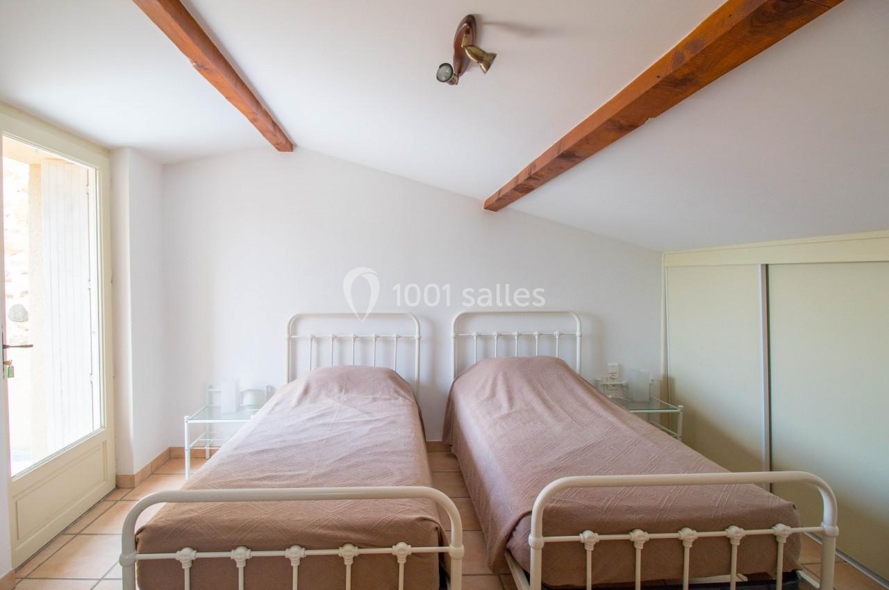 Deux lits simples avec draps roses dans une chambre lumineuse aux poutres apparentes et sol en carrelage.