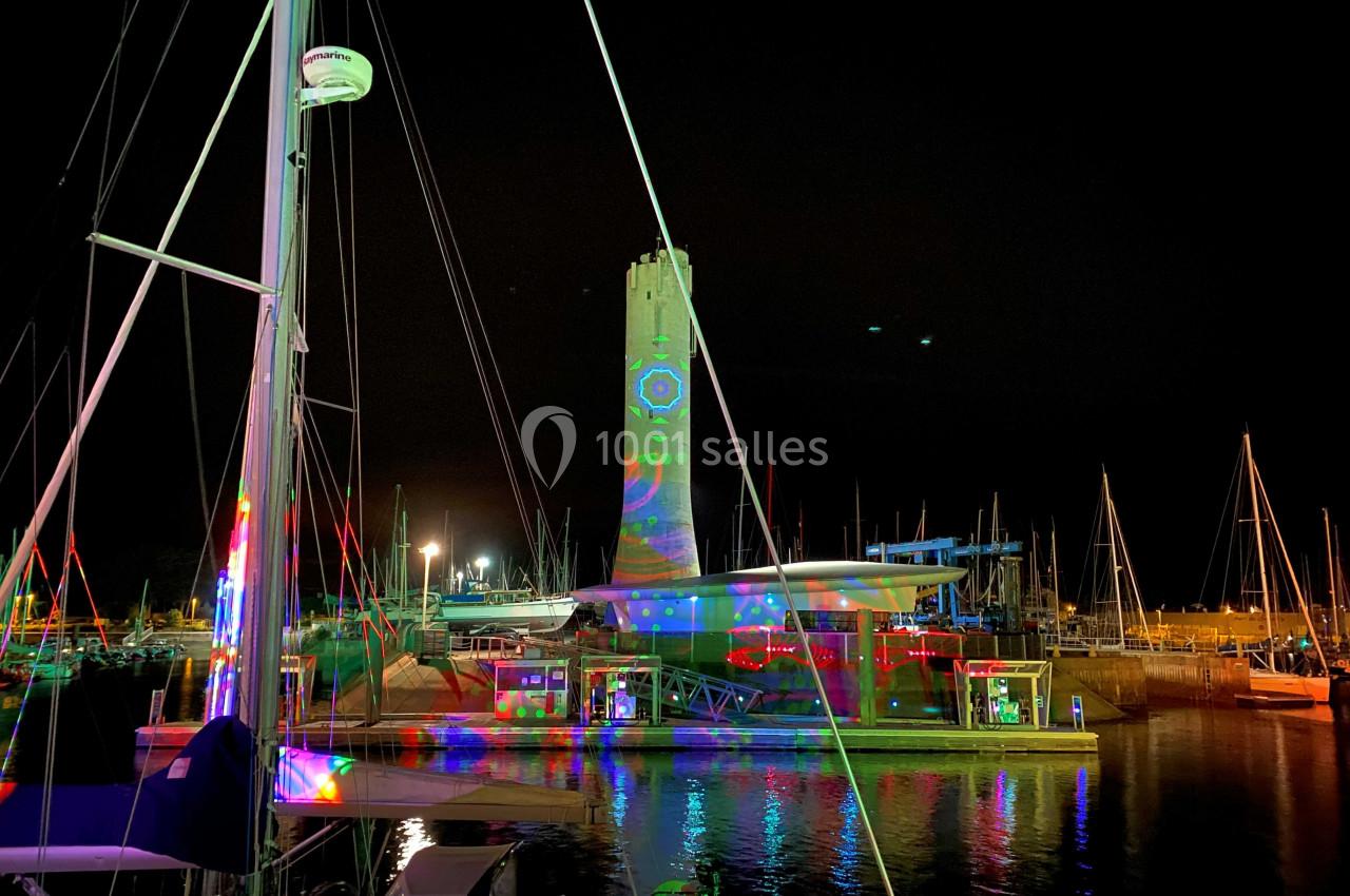 Tour éclairée par des projections colorées au bord d'un port, entourée de voiliers et de reflets sur l'eau la nuit.