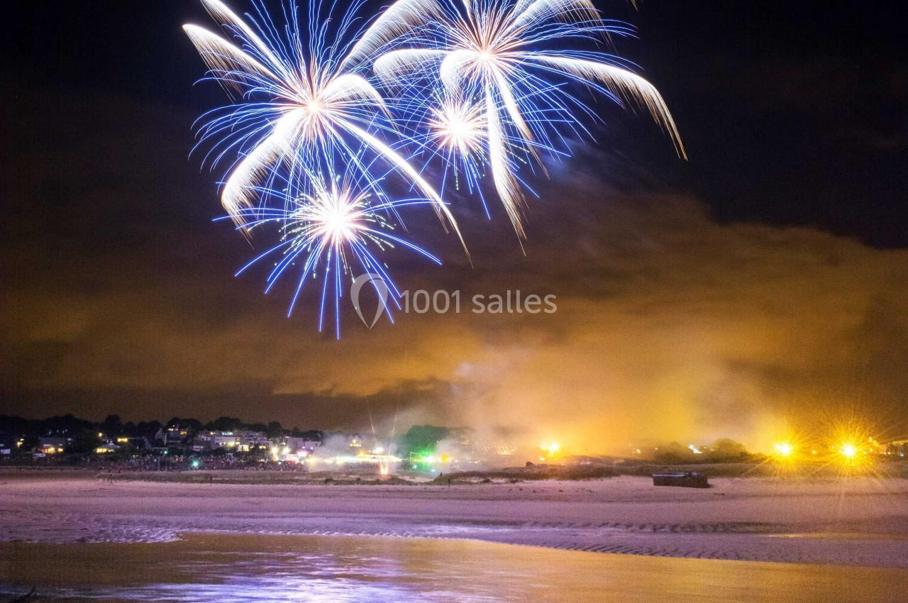 Feux d'artifice colorés illuminant le ciel nocturne au-dessus d'une plage avec des lumières de ville en arrière-plan.