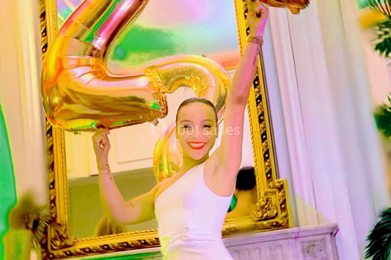 Femme en robe blanche tenant des ballons dorés formant le chiffre 21, devant un miroir orné dans une pièce lumineuse.