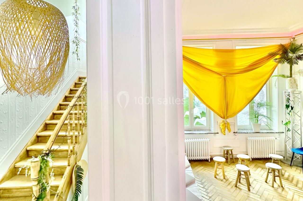 Escalier en bois décoré de plantes à gauche et espace lumineux avec tissu jaune suspendu et tabourets à droite.