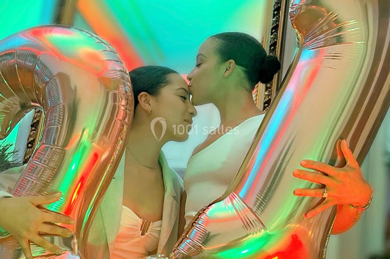 Deux femmes s'enlacent affectueusement devant des ballons métalliques en forme de chiffres, dans un décor coloré.