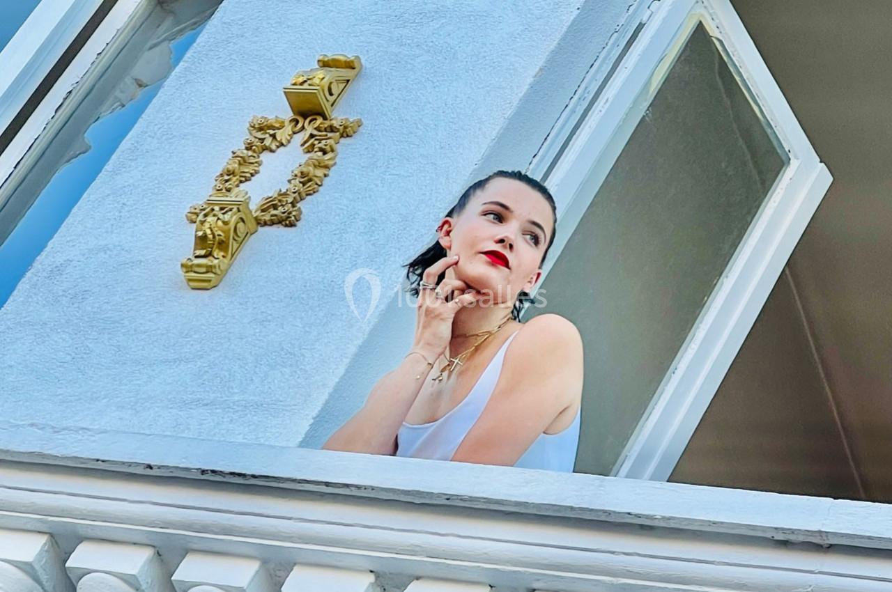 Femme en débardeur blanc regardant par une fenêtre ouverte, appuyée sur le rebord, avec un heurtoir doré visible.