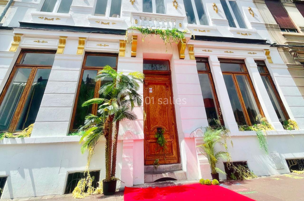 Façade d'un bâtiment blanc avec ornements dorés, porte en bois sculpté, plantes et tapis rouge devant l'entrée.