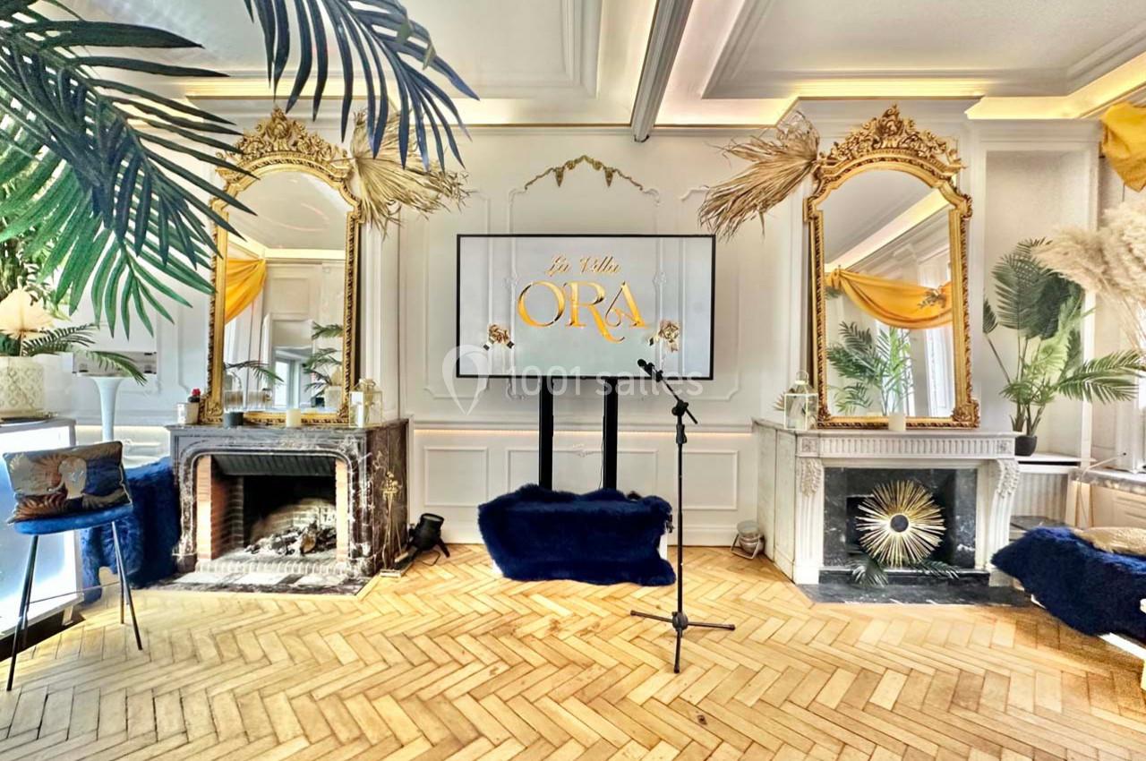 Salle élégante avec parquet en chevrons, miroirs dorés, cheminée décorative et écran affichant ’La Villa Ora’.
