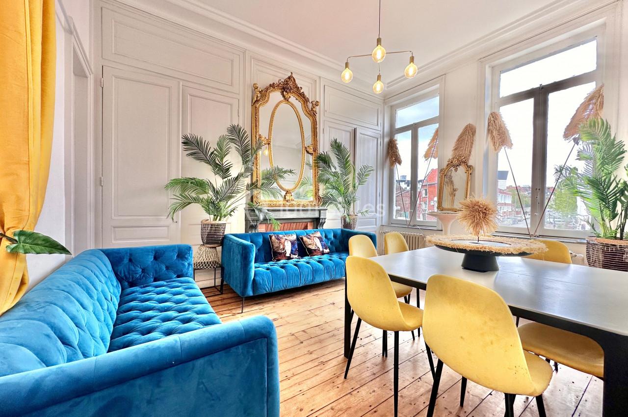 Salon lumineux avec parquet en bois, canapés bleus, chaises jaunes, table noire et miroir doré encadré de plantes.