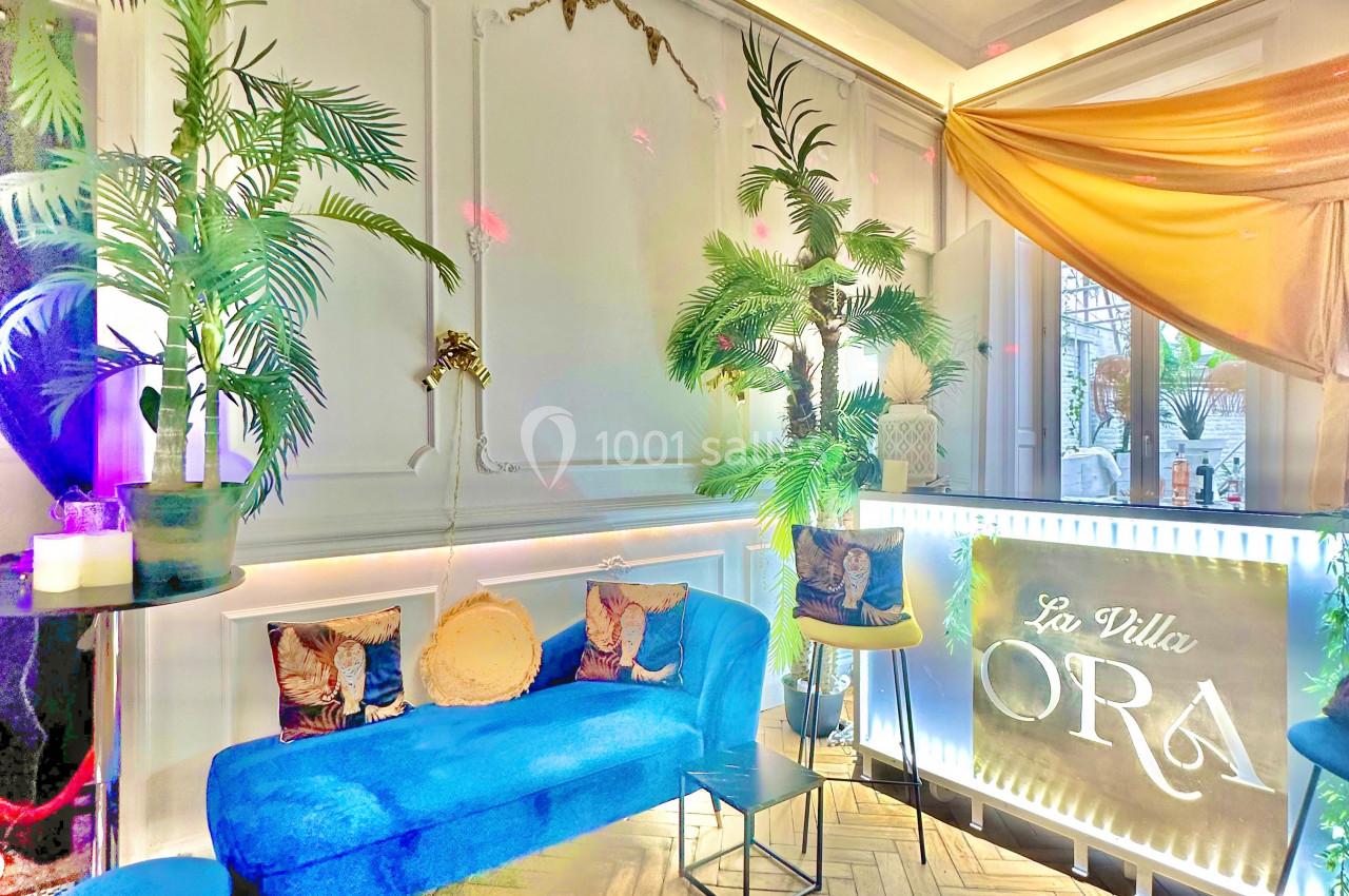 Salon lumineux avec canapé bleu, plantes tropicales, parquet en chevrons et enseigne lumineuse ’La Villa Ora’.