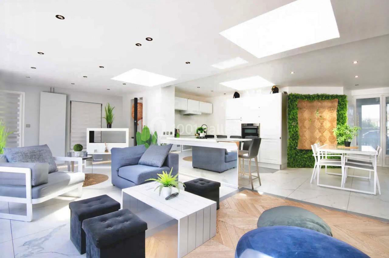 Salon moderne lumineux avec mobilier blanc et bleu, plantes décoratives et mur végétal dans une cuisine ouverte.