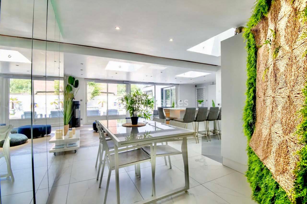 Salle à manger lumineuse avec table en verre, mur végétal, et grandes baies vitrées donnant sur l'extérieur.