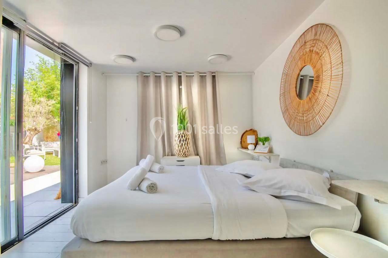 Chambre lumineuse avec lit double, décoration épurée, miroir rond en rotin et vue sur un jardin par une baie vitrée.