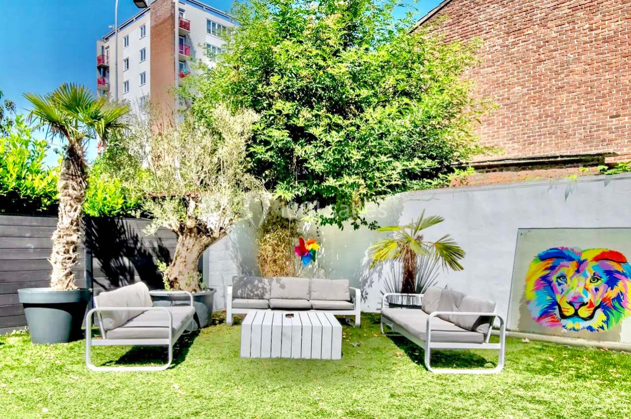 Salon de jardin avec canapés gris, table basse blanche, plantes exotiques et tableau coloré sur un mur extérieur.