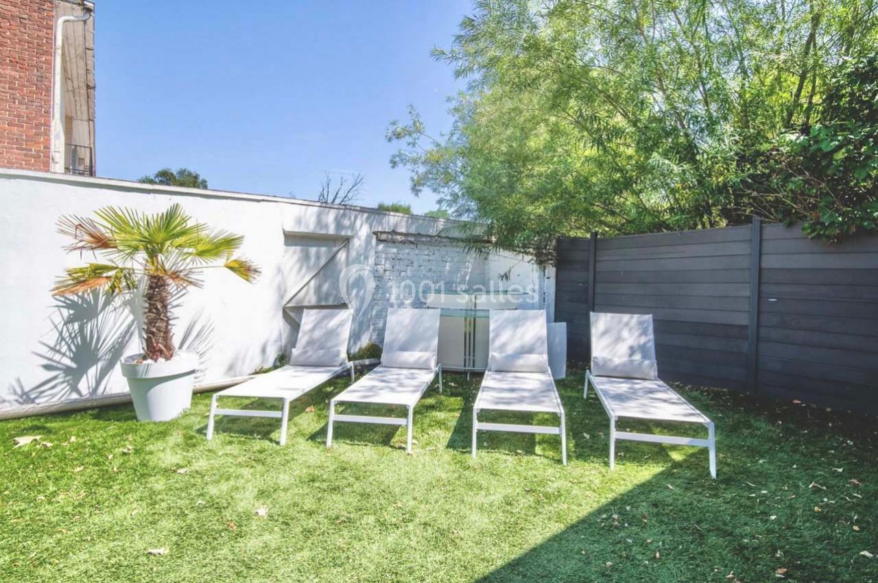 Chaises longues blanches alignées sur une pelouse, près d'un palmier en pot et d'une clôture en bois sombre.