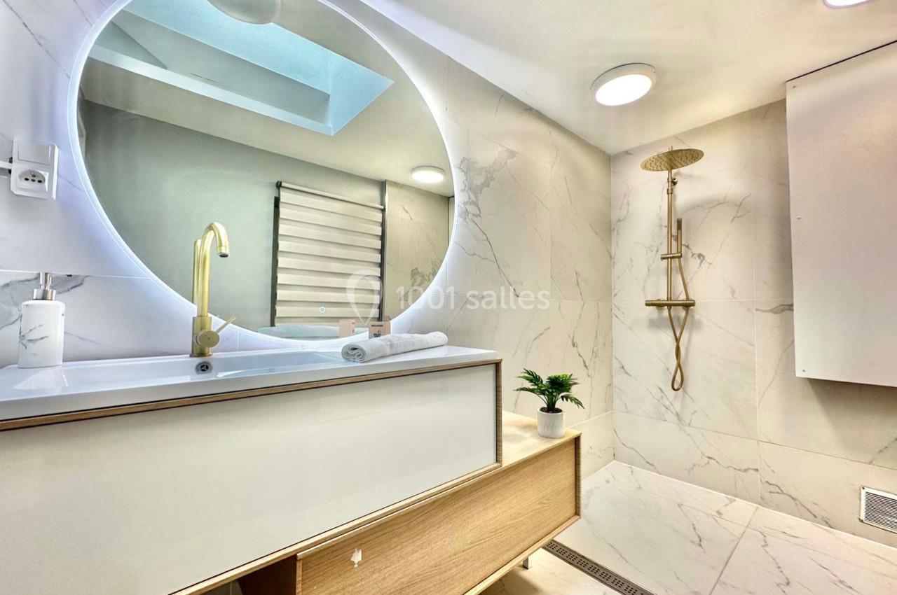 Salle de bain moderne avec lavabo, miroir rond éclairé, douche à effet pluie et murs en marbre clair.