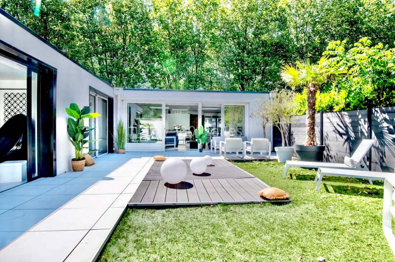 Terrasse moderne avec mobilier design, entourée de verdure et donnant sur une maison aux grandes baies vitrées.