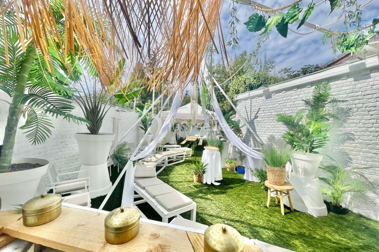 Terrasse extérieure aménagée avec chaises longues, voilages blancs, plantes tropicales et pelouse artificielle.