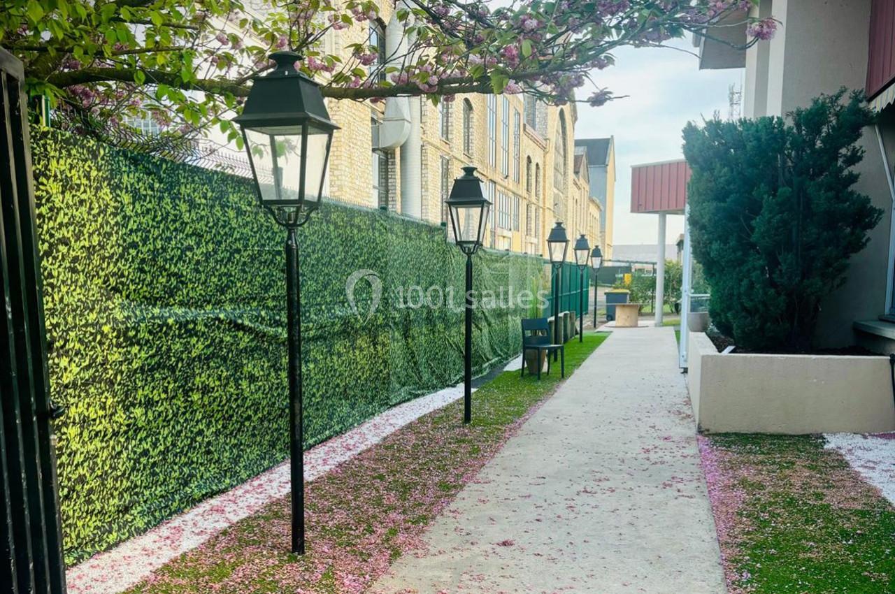 Allée bordée de lampadaires noirs, de haies artificielles et de pétales roses tombés au sol.