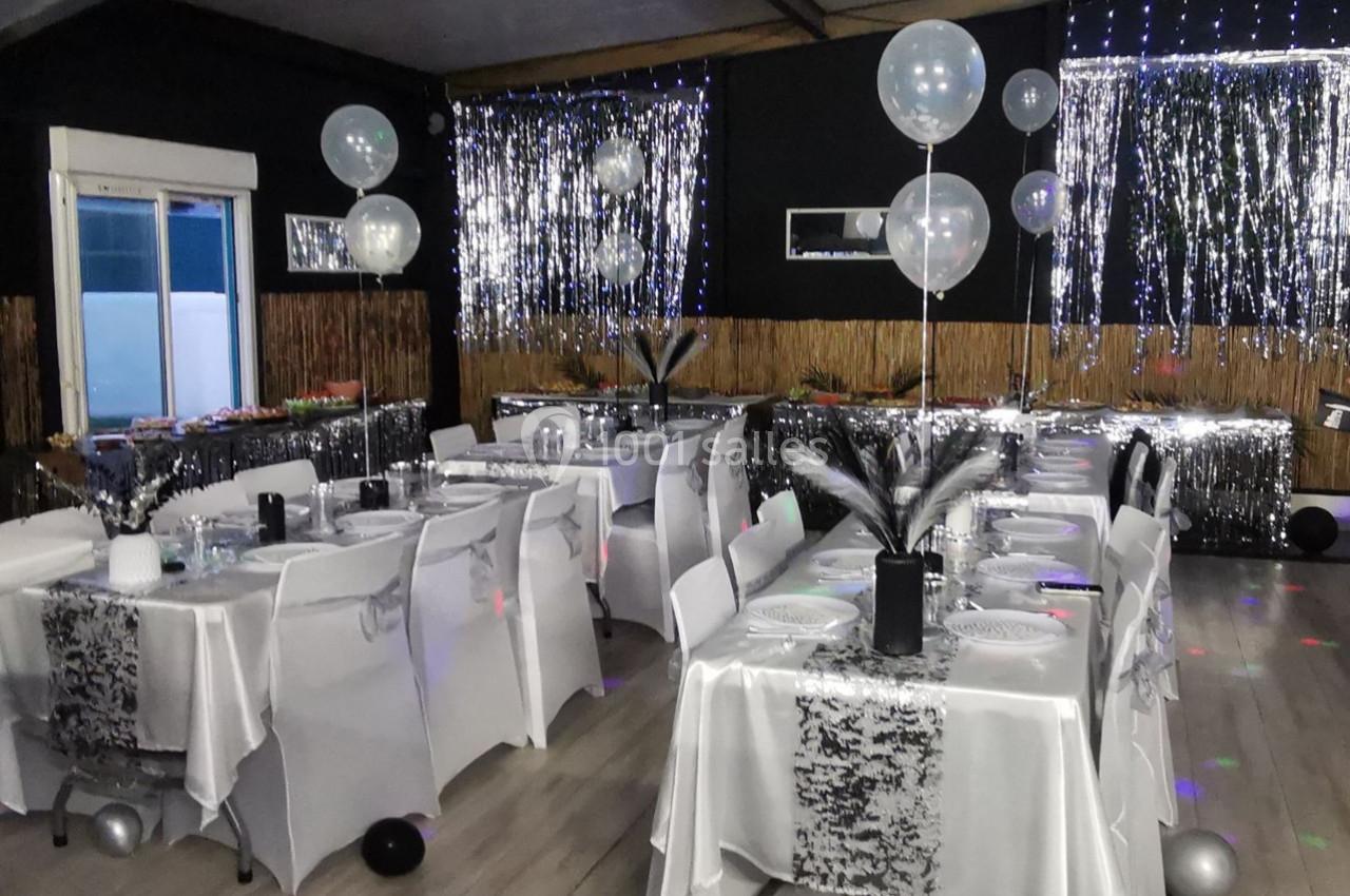 Salle décorée pour une fête avec tables dressées, nappes blanches, ballons transparents et guirlandes argentées.
