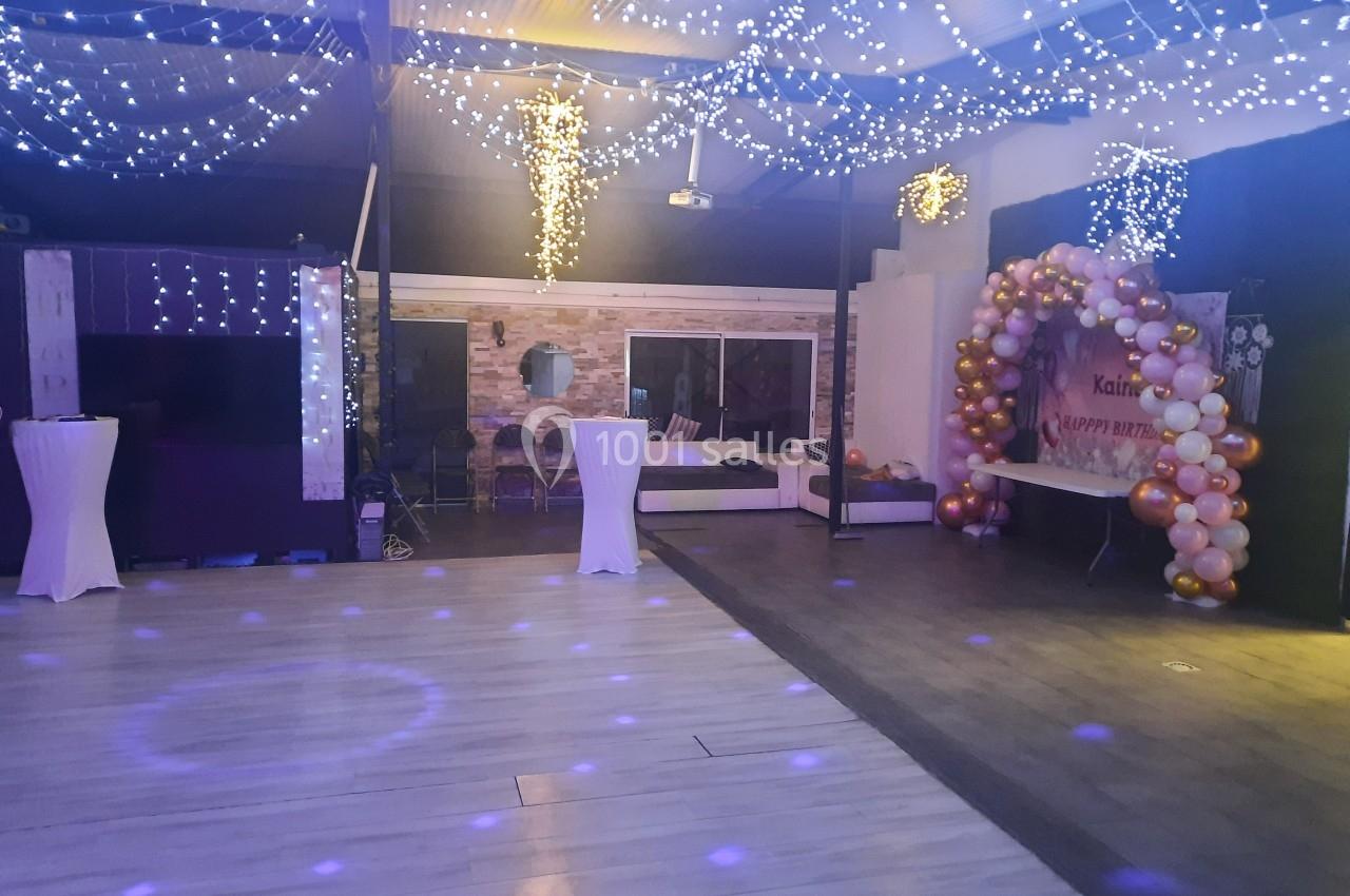 Salle décorée pour une fête avec guirlandes lumineuses, arche de ballons et tables hautes blanches.