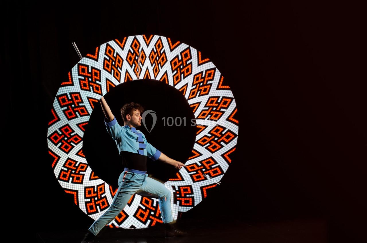 Un artiste en costume bleu exécute une pose dynamique devant un cercle lumineux à motifs colorés.