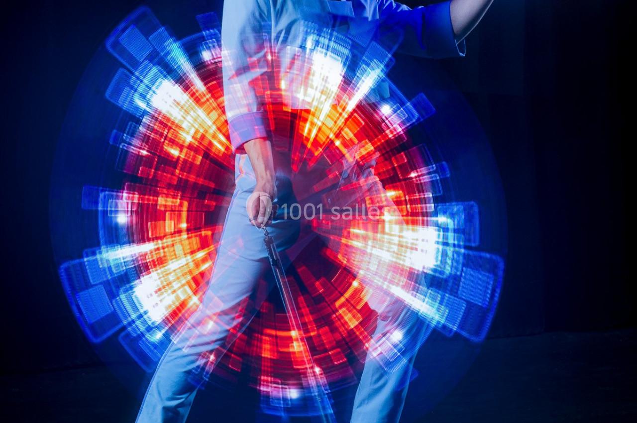 Personne en tenue blanche tenant un objet devant un motif lumineux circulaire rouge et bleu sur fond sombre.