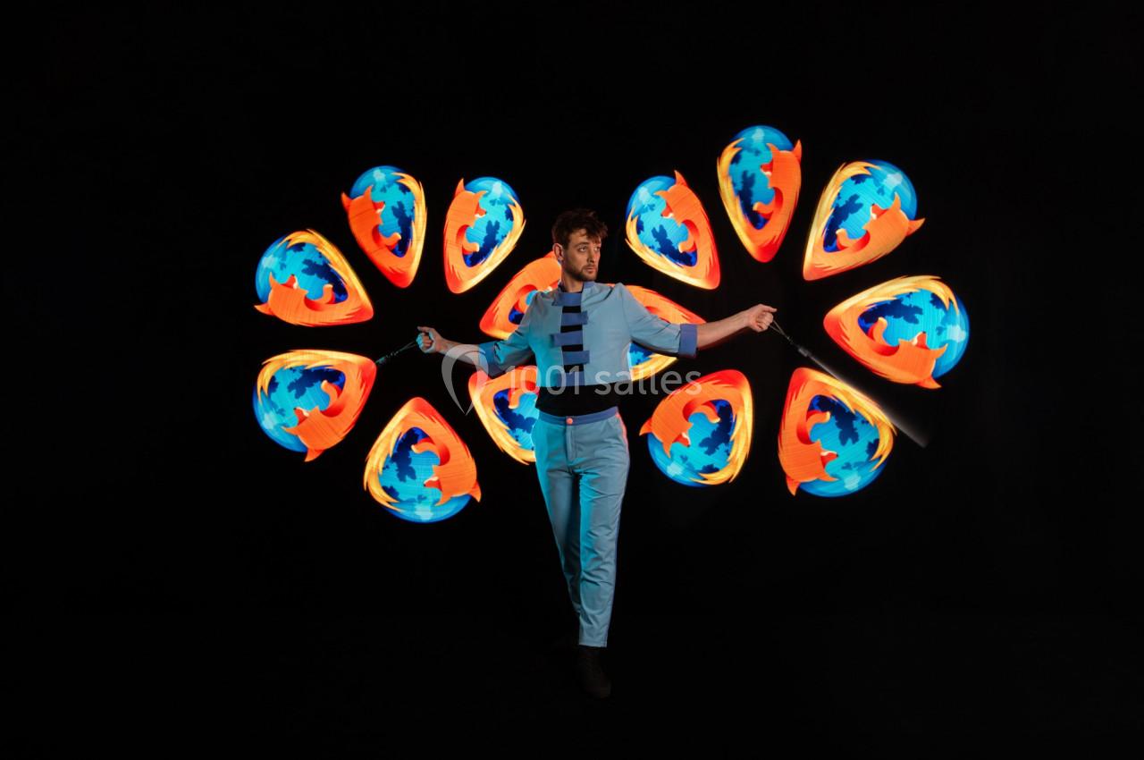 Un homme jongle avec des objets lumineux créant des motifs colorés en mouvement sur un fond noir.
