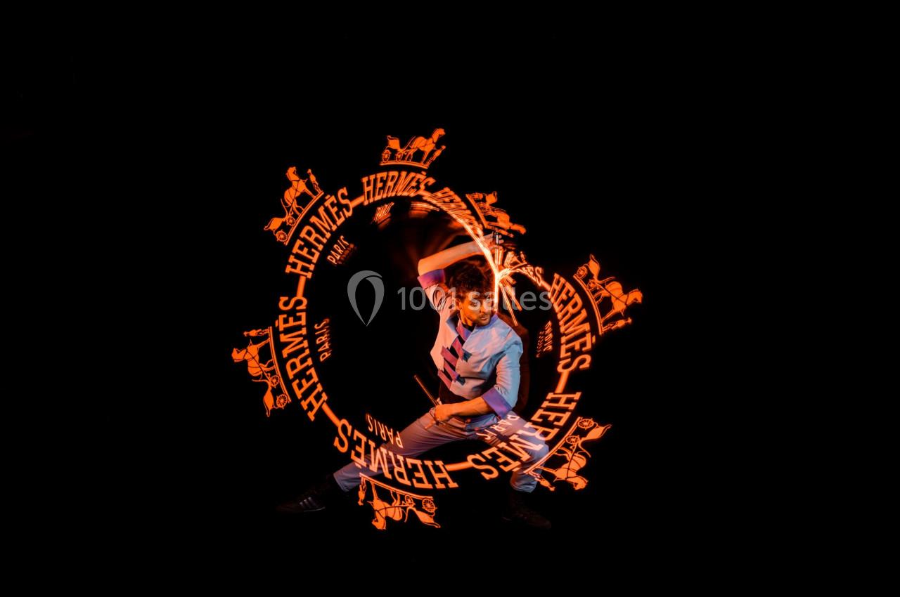 Un homme manipule un bâton lumineux créant des motifs orange avec le logo Hermès sur un fond noir.