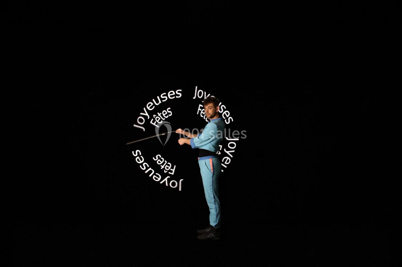 Un homme en tenue bleue manipule un bâton lumineux affichant ’Joyeuses fêtes’ dans un espace sombre.