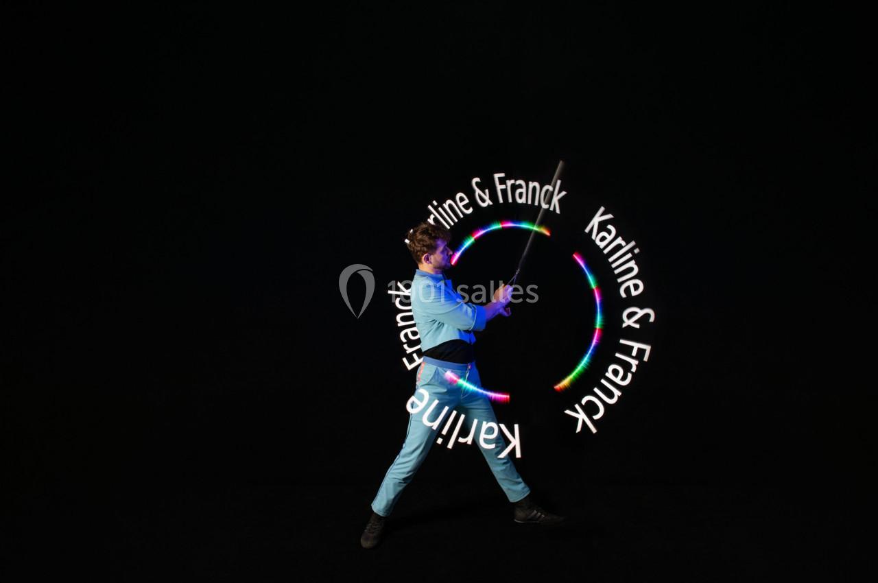 Un jongleur en tenue bleue manipule un bâton lumineux créant des motifs circulaires avec le texte ’Karine & Franck’.
