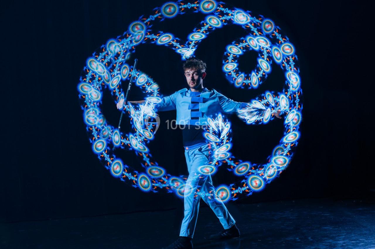 Un jongleur en costume bleu manipule des bâtons lumineux créant des motifs circulaires dans un décor sombre.