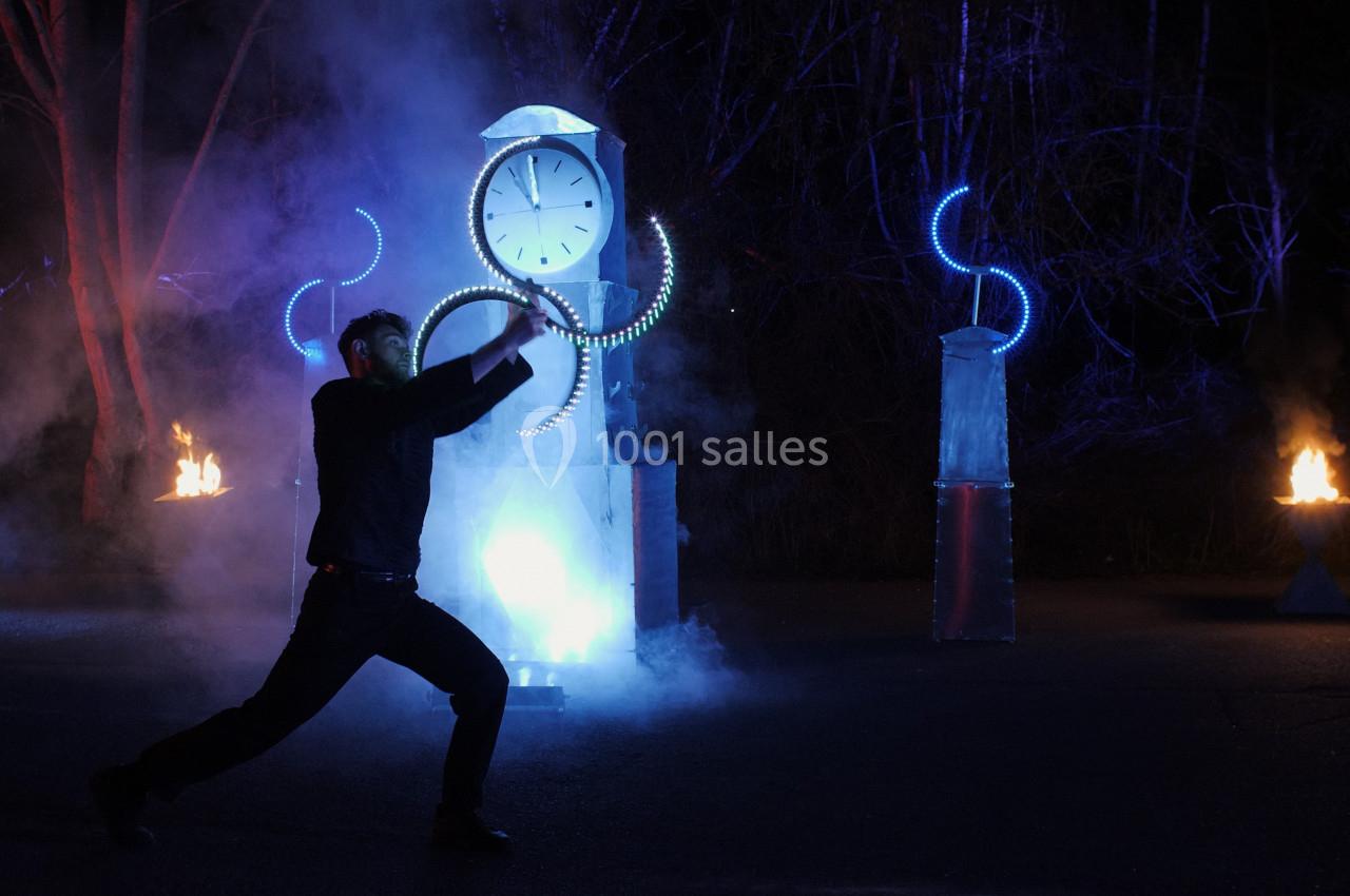 Un homme danse devant une horloge illuminée et des installations lumineuses, entouré de fumée et de flammes.