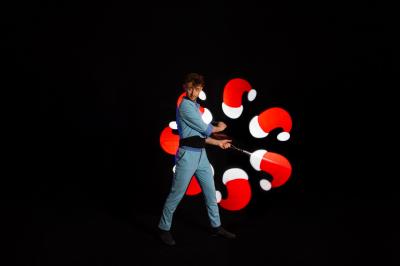 Un jongleur réalise une performance avec des bâtons rouges dans une galerie lumineuse bordée de colonnes.