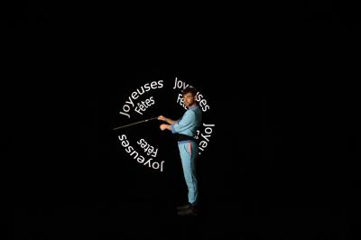 Un jongleur réalise une performance avec des bâtons rouges dans une galerie lumineuse bordée de colonnes.