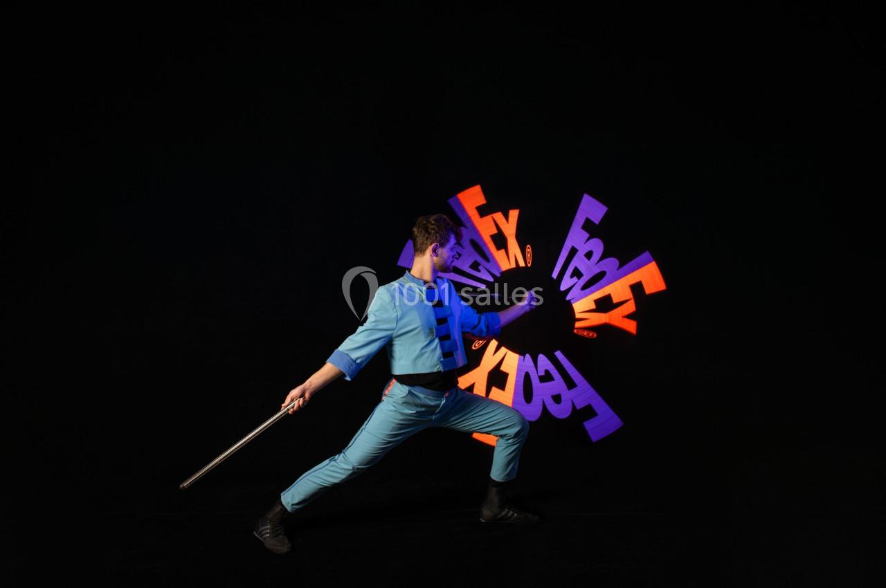 Personne en tenue bleue manipulant un bâton devant des formes lumineuses colorées sur fond noir.