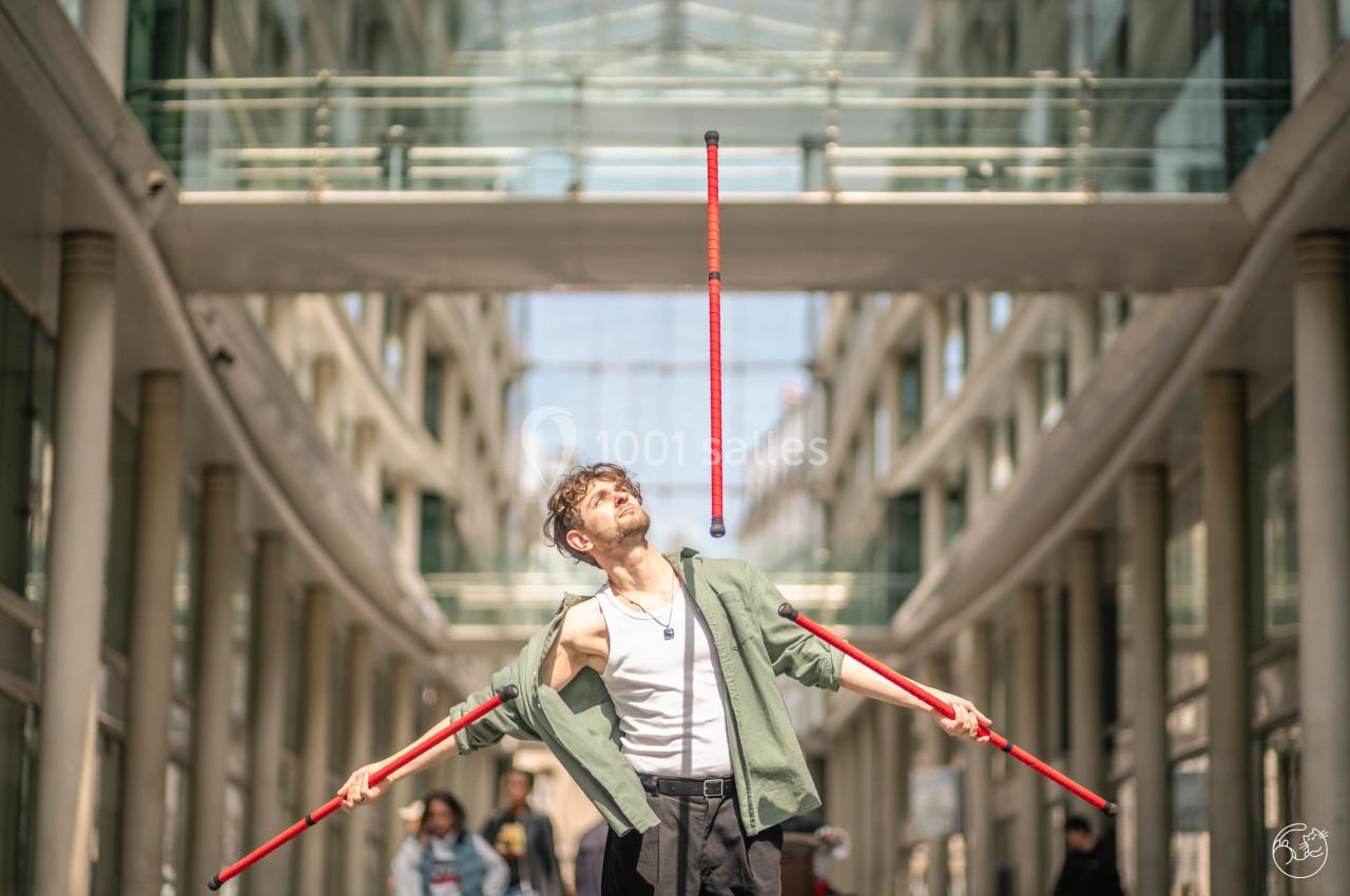 Un jongleur réalise une performance avec des bâtons rouges dans une galerie lumineuse bordée de colonnes.