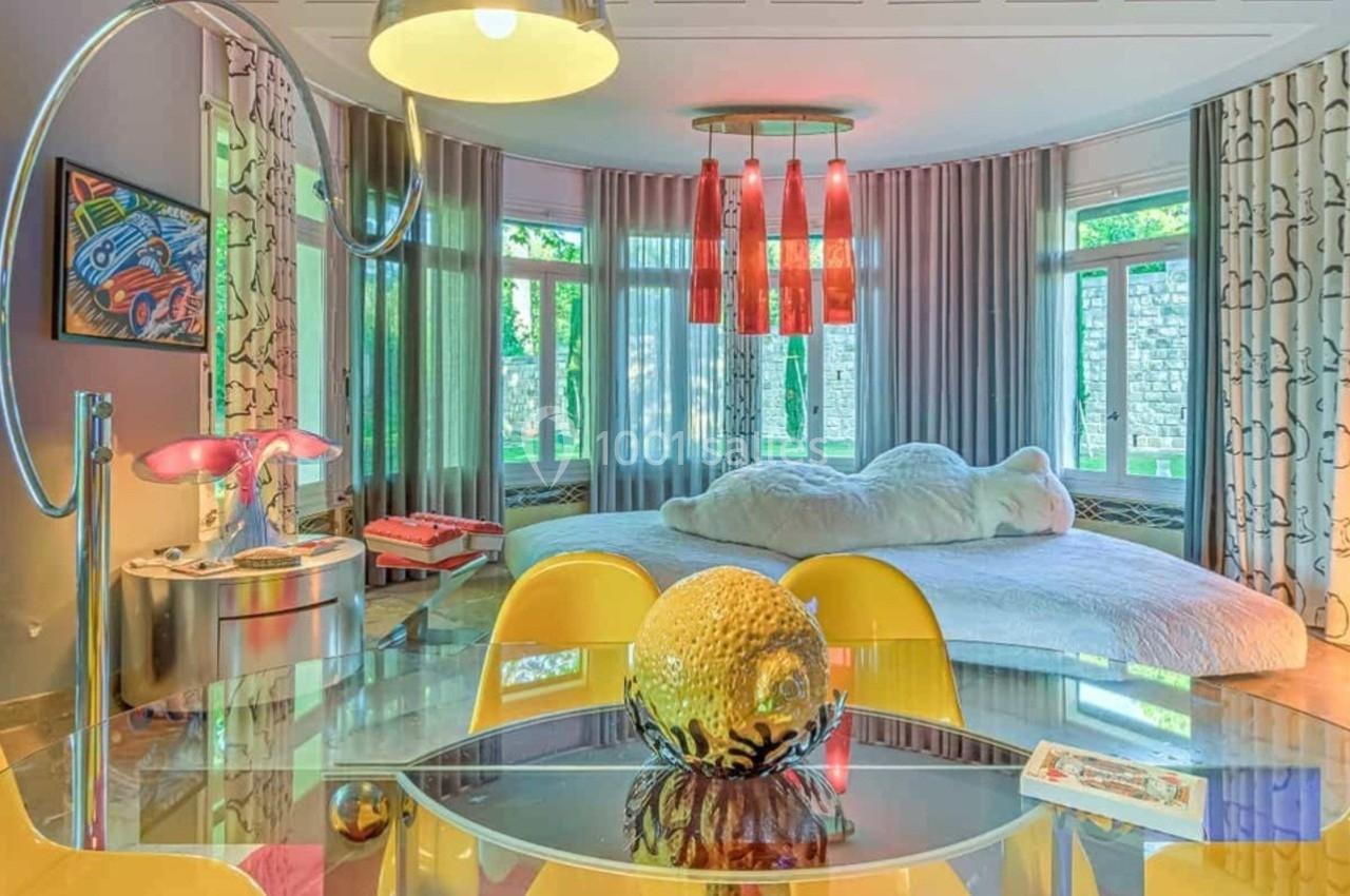 Salle colorée avec table en verre, chaises jaunes, lit rond, rideaux clairs et décorations éclectiques.