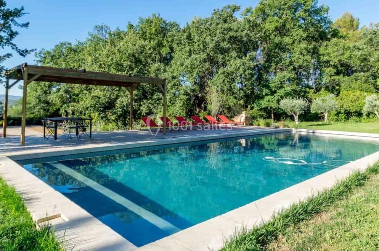 Piscine extérieure entourée de verdure, avec transats rouges et une pergola en bois à proximité.