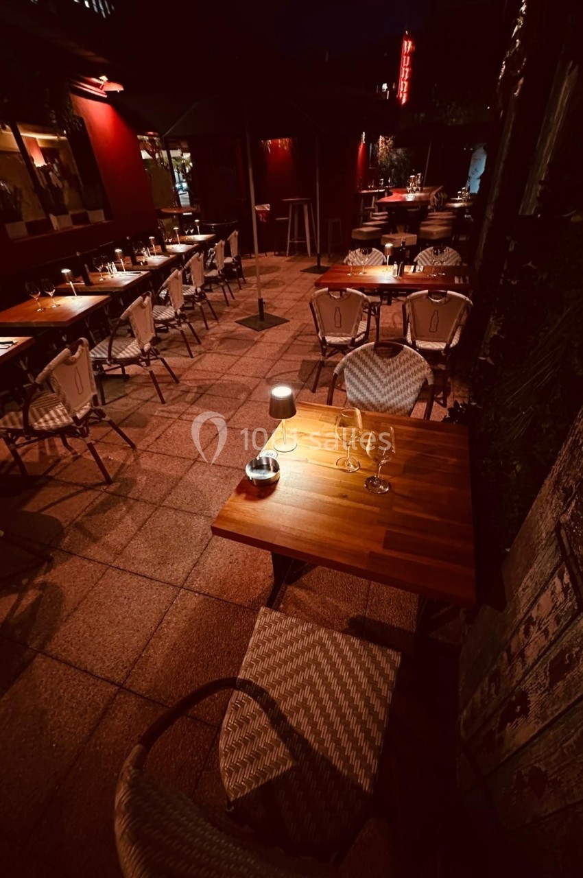 Terrasse d'un restaurant éclairée par des lampes, avec tables en bois et chaises tressées, dans une ambiance nocturne.