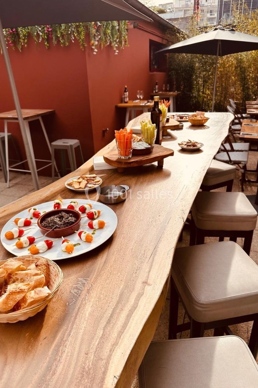 Table en bois avec apéritifs variés, légumes, dips et pain, disposée en terrasse avec chaises et parasols.