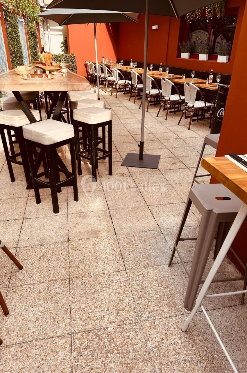 Terrasse extérieure avec tables hautes, chaises et parasols, entourée de murs rouges et de verdure.
