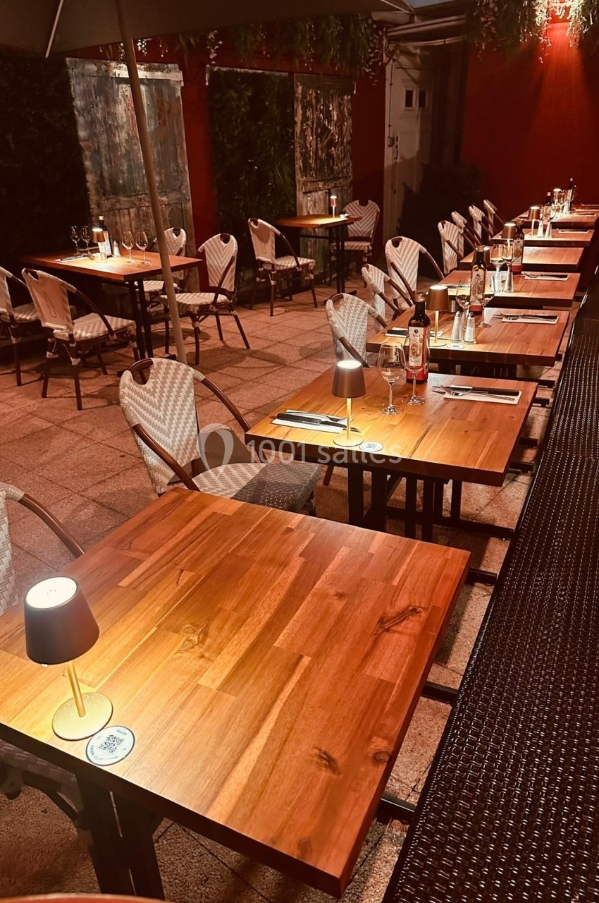 Terrasse de restaurant éclairée en soirée, avec tables en bois, chaises tressées et lampes individuelles.