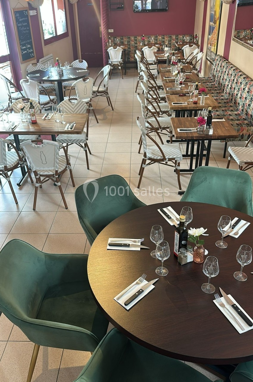 Salle de restaurant lumineuse avec tables dressées, chaises en rotin et fauteuils verts autour d'une table ronde.