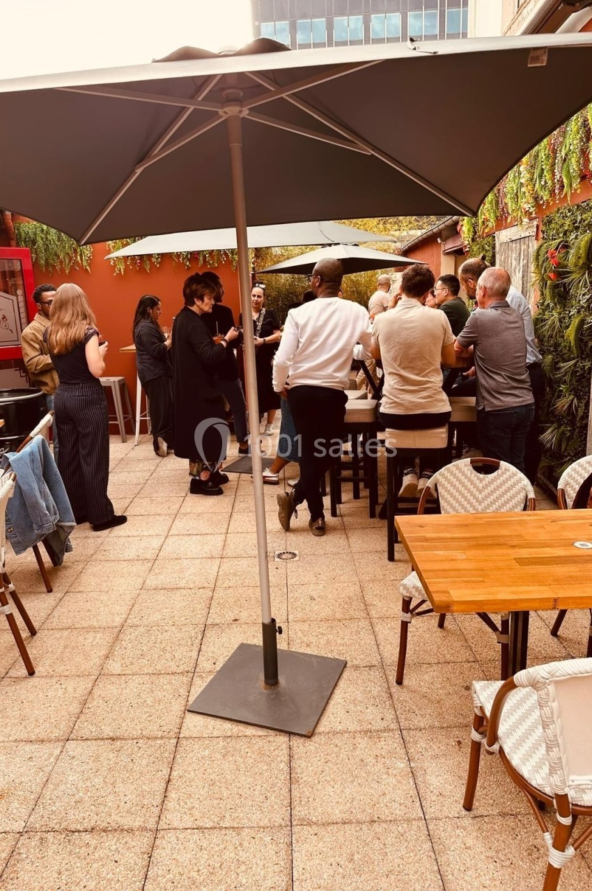 Personnes debout discutant sous un grand parasol sur une terrasse extérieure aménagée avec tables et chaises.